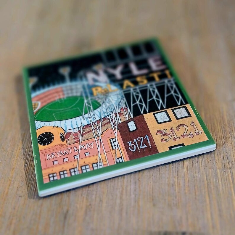 May include: A square ceramic coaster featuring a colourful illustration of a stadium and industrial buildings. The text reads NYLE, PLASTI, BRYANT & MAY, and 3121. The design includes a clock tower, floodlight towers, and a green field under a dark sky.