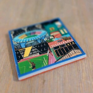 May include: A square ceramic coaster featuring a colorful graphic illustration of a sports stadium with a green field, a blue circular structure, and a yellow number seven. The design includes black geometric shapes and text reading PUNT RD.