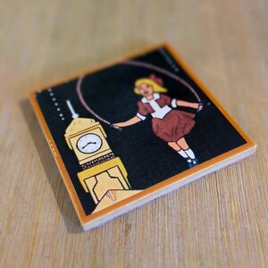 May include: A square ceramic tile featuring a black background with a graphic illustration of a blonde person in a brown dress jumping rope. Next to the person is a yellow clock tower with a white clock face and black hands. The tile has a light orange border.