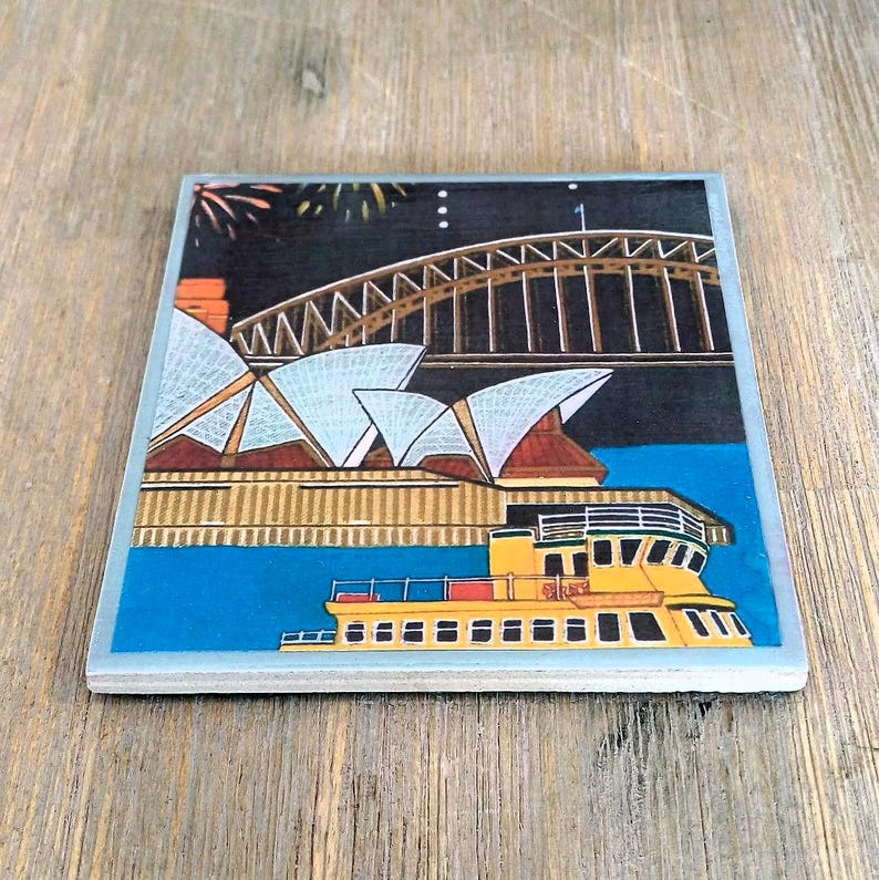 May include: A square ceramic coaster featuring a colorful illustration of the Sydney Opera House and Harbour Bridge at night, with a yellow ferry boat in the foreground. The scene includes a dark sky, blue water, and architectural details.
