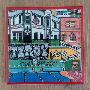 May include: A square ceramic tile with a red border featuring a colorful illustration of a street scene. The image depicts the Napier Hotel, Prince of Prussia, street signs for Johnson St and Brunswick St, a Fitzroy sign, and a person with a beard.
