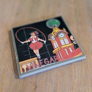 May include: A square ceramic coaster featuring a black background with a graphic illustration of a person jumping rope next to a red and yellow clock tower. The word VINEGAR is printed in large, bold, light-coloured letters at the bottom.