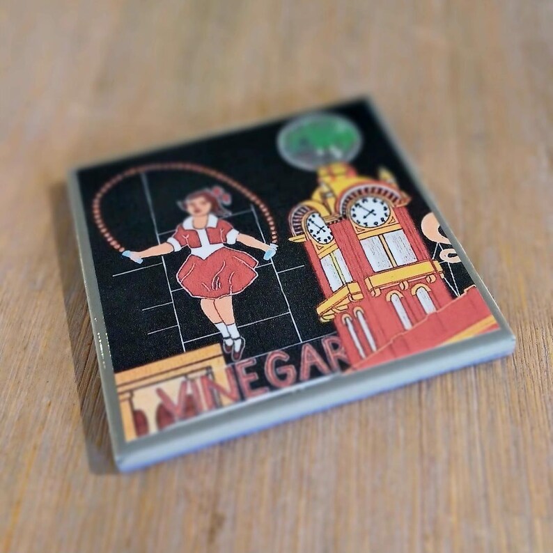 May include: A square ceramic coaster featuring a black background with a graphic illustration of a person jumping rope next to a red and yellow clock tower. The word VINEGAR is printed in large, bold, light-coloured letters at the bottom.
