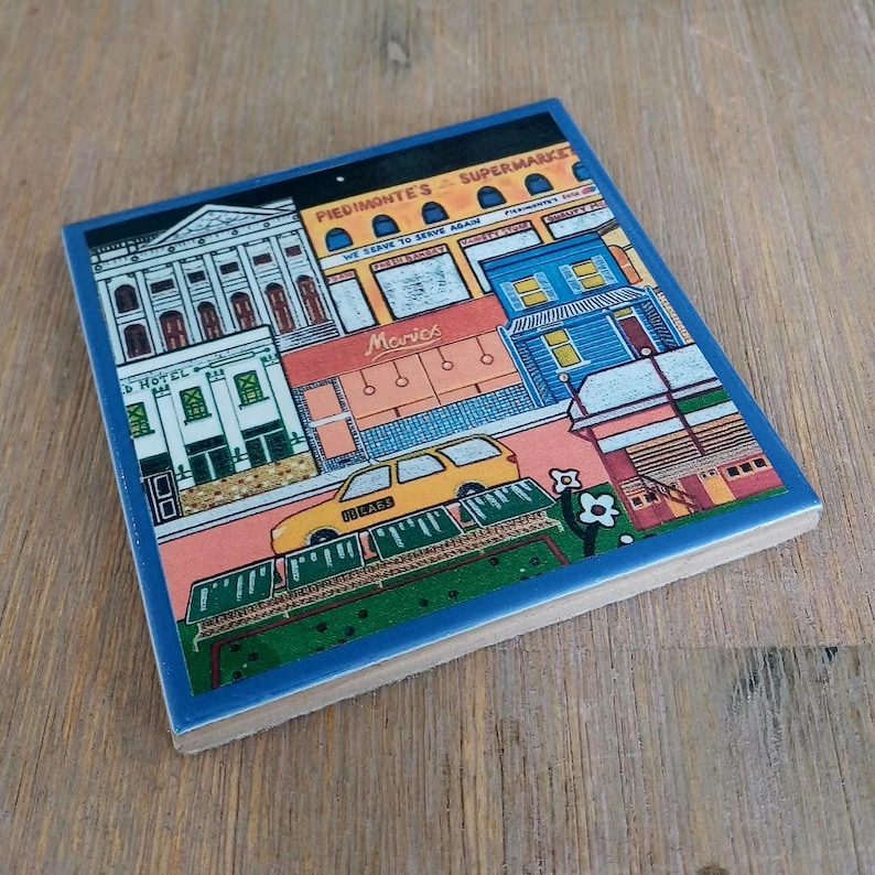 May include: A square ceramic tile featuring a colorful illustration of a city street scene. The artwork depicts a white hotel, a yellow taxi, and storefronts labeled Piedmont's Supermarket and Marios. The design includes blue, orange, and green tones.