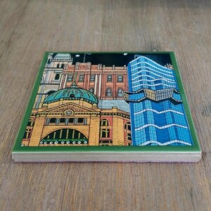 Op de afbeelding: Een vierkante keramische onderzetter met een kleurrijke illustratie van een stadssilhouet. Het ontwerp bevat een geel gebouw met een groene koepel en een hoge blauwe glazen wolkenkrabber tegen een nachtelijke hemel, omlijst door een donkergroene rand.