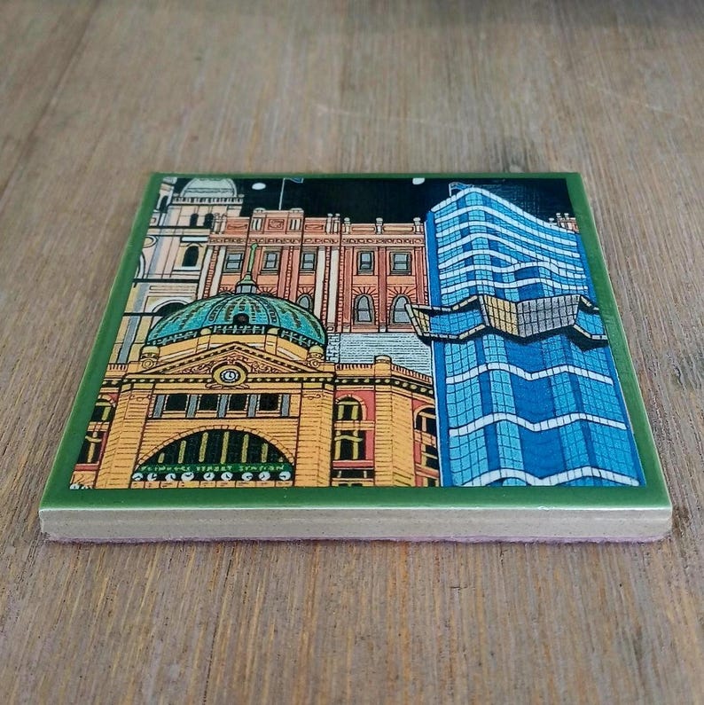 Op de afbeelding: Een vierkante keramische onderzetter met een kleurrijke illustratie van een stadssilhouet. Het ontwerp bevat een geel gebouw met een groene koepel en een hoge blauwe glazen wolkenkrabber tegen een nachtelijke hemel, omlijst door een donkergroene rand.