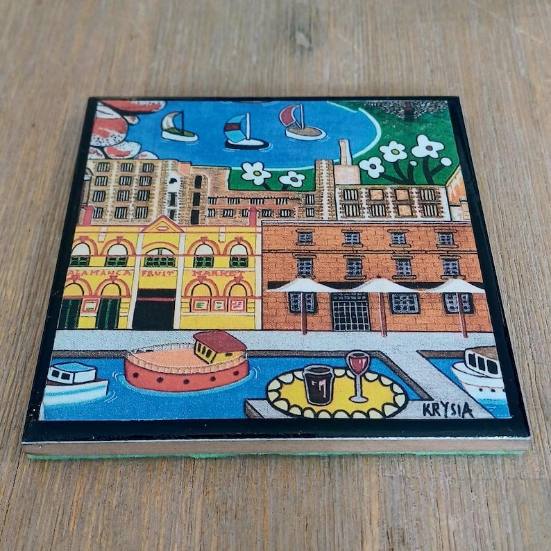 Op de afbeelding: Een vierkante onderzetter met een kleurrijke illustratie van een waterkant met gebouwen, waaronder de Salamanca Fruit Market, boten op blauw water en een tafel met een glas en beker. De kunstenaarshandtekening Krysia staat in de hoek.