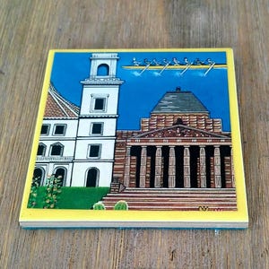 Puede incluir: Un azulejo cuadrado de cer&aacute;mica con una ilustraci&oacute;n colorida de un edificio blanco con una torre alta junto a una estructura marr&oacute;n tipo templo con columnas. Sobre los edificios, se representa un equipo de remo en un bote bajo un cielo azul.