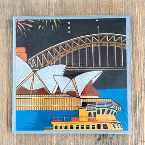 May include: A square coaster featuring a colorful illustration of the Sydney Opera House and Harbour Bridge at night. The scene includes a yellow ferry on blue water, white architectural sails, a dark sky with fireworks, and a light gray border.