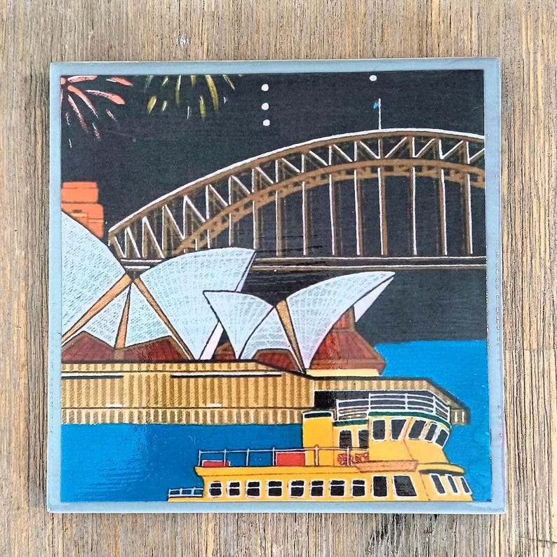 May include: A square coaster featuring a colorful illustration of the Sydney Opera House and Harbour Bridge at night. The scene includes a yellow ferry on blue water, white architectural sails, a dark sky with fireworks, and a light gray border.