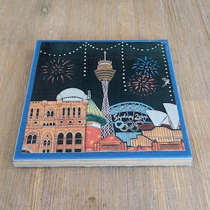May include: A square ceramic coaster featuring a nighttime illustration of the Sydney skyline with the Sydney Tower, the Opera House, and the Olympic rings. The text Sydney 2000 is printed in white, set against a black sky with colorful fireworks.