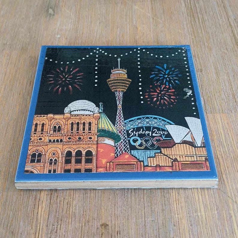 May include: A square ceramic coaster featuring a nighttime illustration of the Sydney skyline with the Sydney Tower, the Opera House, and the Olympic rings. The text Sydney 2000 is printed in white, set against a black sky with colorful fireworks.