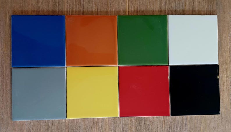 May include: A collection of eight square tiles arranged in two rows of four. The tiles feature a smooth, glossy finish in distinct solid colors including blue, orange, green, white, gray, yellow, red, and black, all displayed in a grid layout.