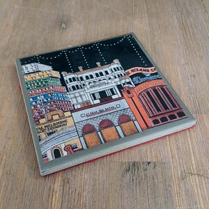 May include: A square coaster featuring a colorful illustration of St Kilda landmarks, including the Hotel Esplanade, St Kilda Sea Baths, and the Palais Theatre. The design includes text labels for each building against a dark night sky background.