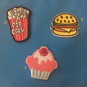 Może przedstawiać: Trzy haftowane naszywki do prasowania: czerwony i biały kubełek popcornu z napisem "POP CORN", żółty hamburger z sałatą, pomidorem i serem oraz różowy cupcake z białym lukrem i wiśnią na wierzchu.