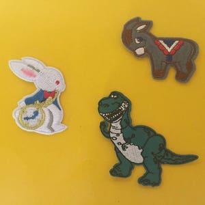 Puede incluir: Tres parches bordados para planchar: un conejo blanco con un reloj de bolsillo, un dinosaurio verde y un burro gris con una silla de montar roja, blanca y azul.