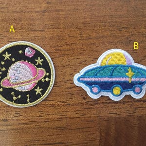 May include: Two embroidered iron-on patches, one with a pink planet and stars on a black background, and the other with a blue and green spaceship on a white background.