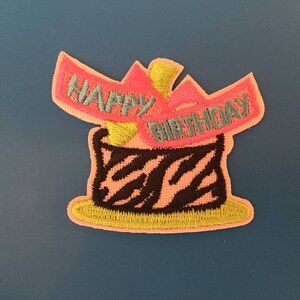 Puede incluir: Un parche bordado de color rosa y negro con las palabras "Happy Birthday" y un diseño de pastel.
