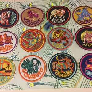 Op de afbeelding: Twaalf geborduurde strijklabels met de astrologische dierenriemtekens. Elk label is rond en heeft een ander sterrenbeeld met de naam eronder. De labels zijn in een rasterpatroon op een groene en bruine achtergrond gerangschikt.