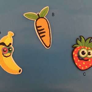 Peut inclure: Trois patchs brodés thermocollants représentant une banane, une carotte et une fraise avec des visages de dessin animé. La banane est jaune avec des yeux noirs et une bouche rouge. La carotte est orange avec des yeux noirs et une bouche noire. La fraise est rouge avec des yeux noirs et une bouche noire.