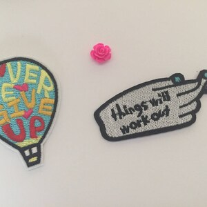 Könnte beinhalten: Zwei gestickte Bügelbilder. Eines ist blau, gelb und rot mit dem Text "Never Give Up" in einer Heißluftballonform. Das andere ist grau mit dem Text "Things will work out" in einer Handform.