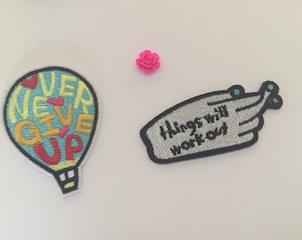 Könnte beinhalten: Zwei gestickte Bügelbilder. Eines ist blau, gelb und rot mit dem Text "Never Give Up" in einer Heißluftballonform. Das andere ist grau mit dem Text "Things will work out" in einer Handform.