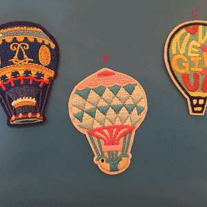 May include: Three embroidered iron-on patches shaped like hot air balloons. One is blue with gold accents and a crown, one is blue, white, and pink with a geometric pattern, and one is yellow with the words "Never Give Up" in red and pink.