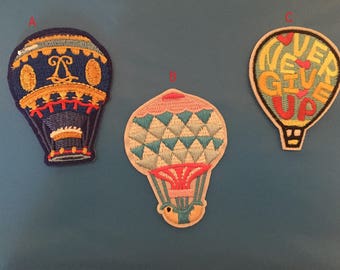Könnte beinhalten: Drei gestickte Bügelbilder in Form von Heißluftballons. Einer ist blau mit goldenen Akzenten und einer Krone, einer ist blau, weiß und rosa mit einem geometrischen Muster und einer ist gelb mit den Worten "Never Give Up" in rot und rosa.