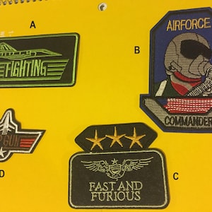May include: Four embroidered patches with different designs. The first patch is green and black with a fighter jet and the words "FIGHTING". The second patch is blue, red, white, and grey with a pilot wearing a helmet and the words "AIRFORCE COMMANDER". The third patch is black with three gold stars and the words "FAST AND FURIOUS". The fourth patch is brown, red, and white with a fighter jet and the words "TOP GUN".