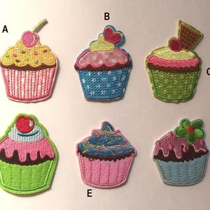 May include: Ten colorful embroidered iron-on cupcake patches. The patches are in various colors, including pink, blue, green, yellow, and brown. Each patch features a different cupcake design with frosting, sprinkles, and cherries.