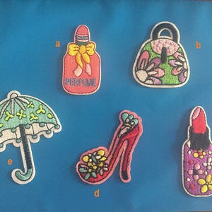 Puede incluir: Cinco parches bordados para planchar que representan una botella de perfume, un bolso de mano, un paraguas, un zapato de tacón alto y un tubo de lápiz labial. Los parches son coloridos y presentan diseños florales.