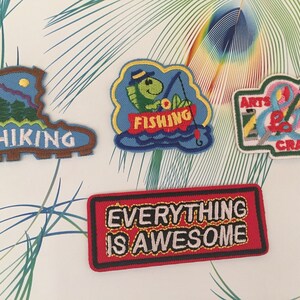 May include: Five embroidered patches with various themes. The patches are blue, brown, green, red, and white. The patches feature the words "Hiking", "Fishing", "Arts & Crafts", and "Everything is Awesome".