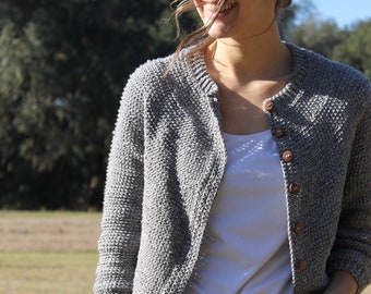 Op de afbeelding: Een vrouw draagt een grijze gebreide cardigan met bruine knopen, een wit T-shirt en een blauwe jeans.
