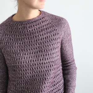 Puede incluir: Un suéter de punto morado con cuello redondo y mangas largas. El suéter tiene un patrón de punto texturizado.