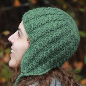 May include: A green crocheted hat with ear flaps. The hat is worn by a person with long brown hair. The person is wearing a blue denim jacket.