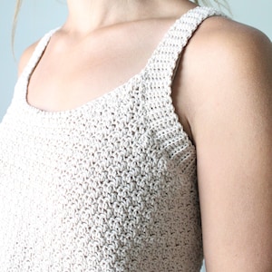 May include: A close-up of a crocheted tank top in a light beige color. The top has a simple, square neckline and thin straps.