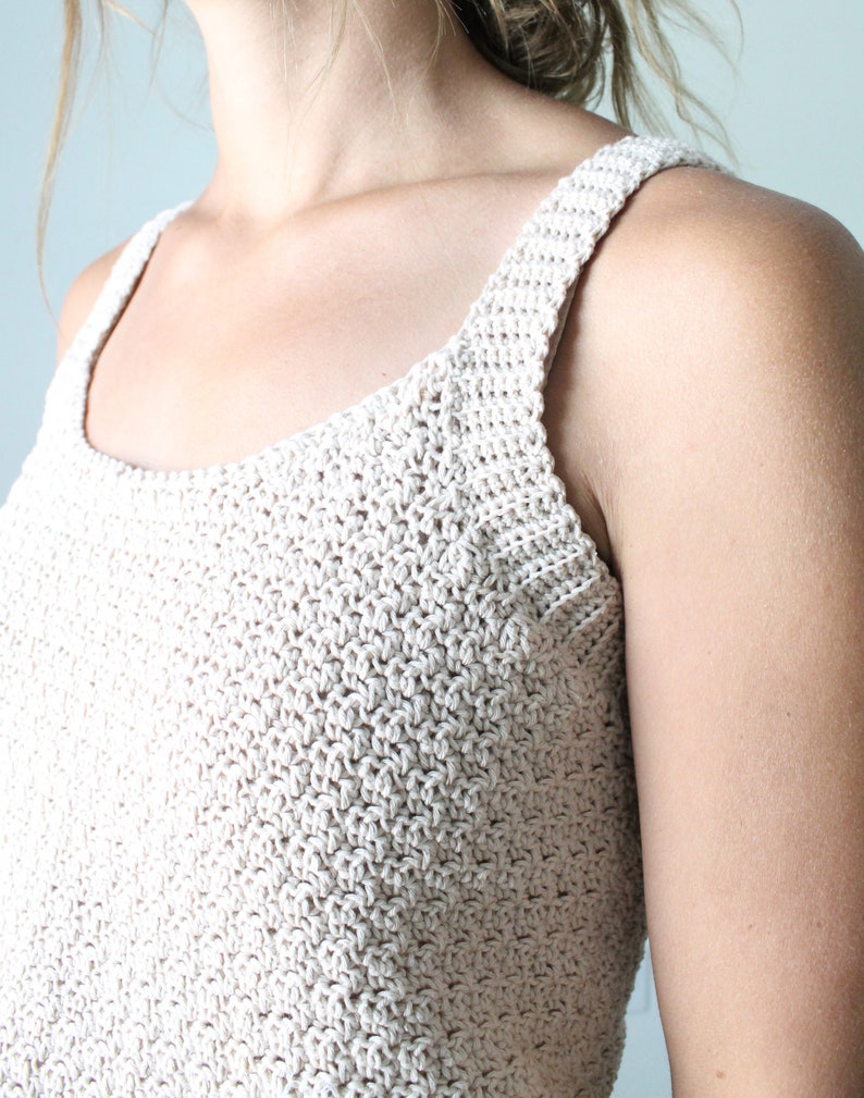 May include: A close-up of a crocheted tank top in a light beige color. The top has a simple, square neckline and thin straps.