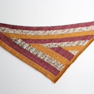 Pode incluir: Um xale triangular com um padrão de listras em tons de bordô, amarelo mostarda e creme. O xale é feito de crochê e tem uma borda franjada.