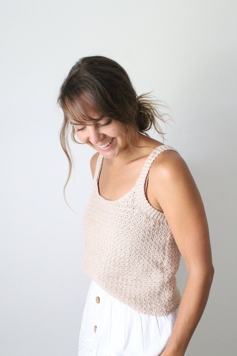 May include: A woman wearing a light brown crocheted tank top and a white skirt with buttons. She is smiling and looking down.
