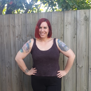 May include: A woman wearing a dark purple crochet tank top with a scoop neck. She is standing in front of a wooden fence.