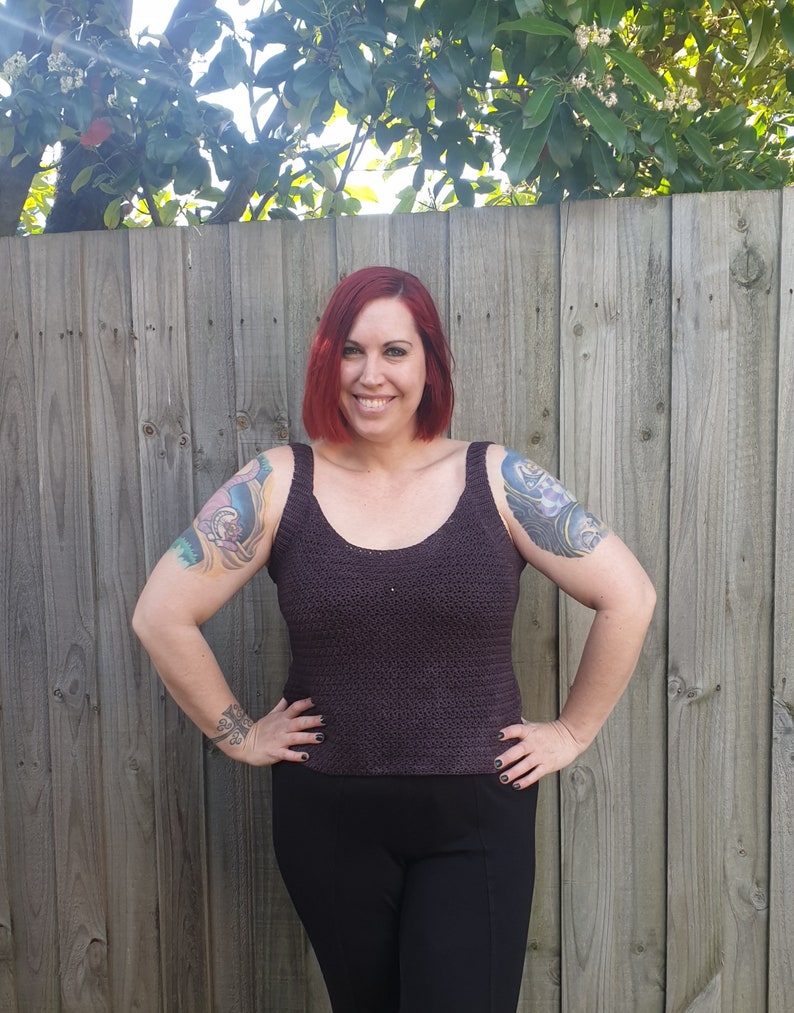 May include: A woman wearing a dark purple crochet tank top with a scoop neck. She is standing in front of a wooden fence.