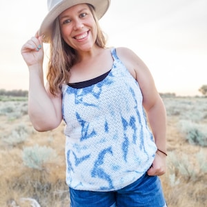 May include: A woman wearing a white and blue tie-dye tank top with a white hat and blue denim shorts. She is standing in a field of dry grass.