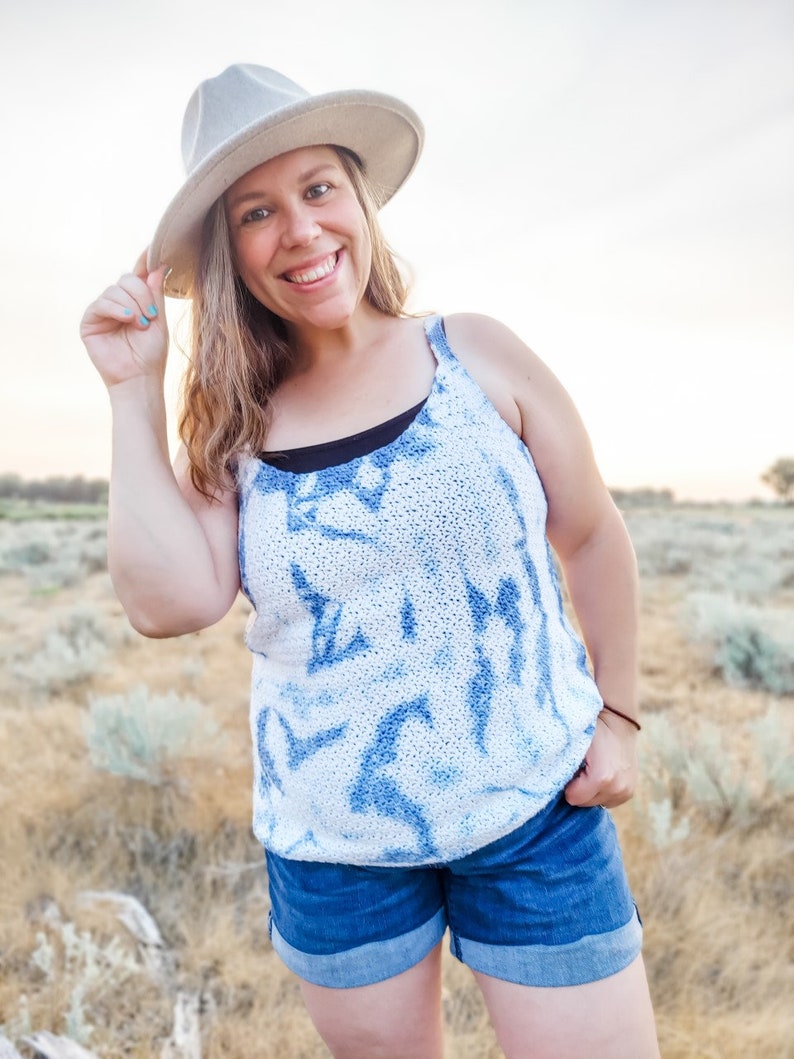 May include: A woman wearing a white and blue tie-dye tank top with a white hat and blue denim shorts. She is standing in a field of dry grass.