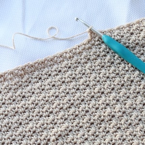 May include: A close-up of a beige crocheted fabric with a teal crochet hook. The fabric is being worked on with a white yarn.