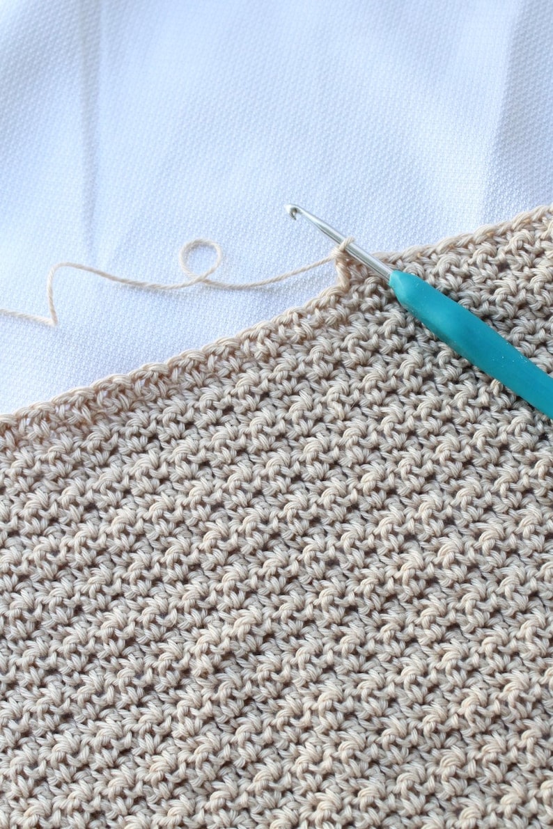 May include: A close-up of a beige crocheted fabric with a teal crochet hook. The fabric is being worked on with a white yarn.