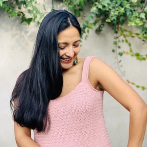 May include: A woman wearing a pink crocheted tank top with a scoop neck. She is smiling and looking down.