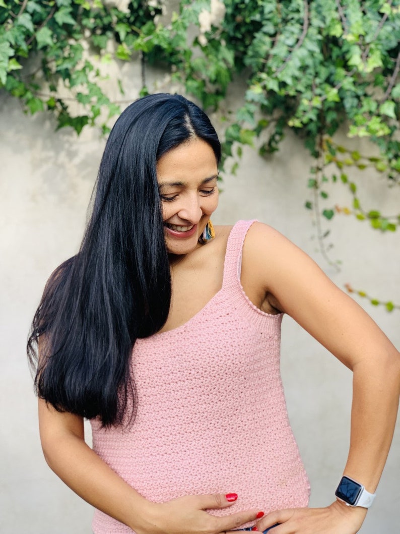 May include: A woman wearing a pink crocheted tank top with a scoop neck. She is smiling and looking down.