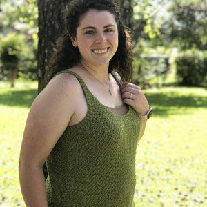 May include: A woman wearing a green sleeveless crocheted top with a scoop neck. She is standing in front of a tree and smiling at the camera.