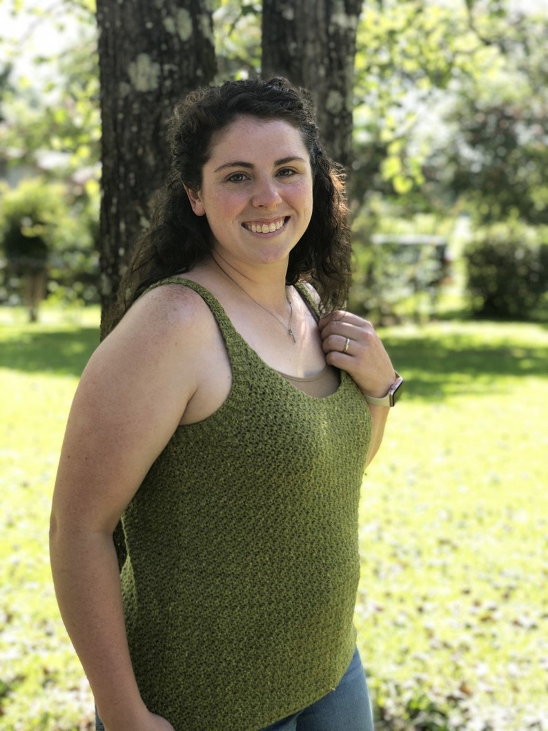 May include: A woman wearing a green sleeveless crocheted top with a scoop neck. She is standing in front of a tree and smiling at the camera.