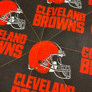 May include: Close-up of a fabric featuring the Cleveland Browns football team logo. The design includes repeating orange helmets and the team name in orange block letters on a black background. Gold stitching is visible.